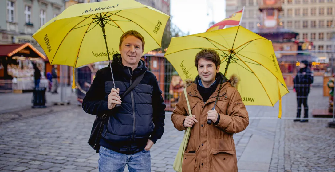 Two men holding yellow Walkative! umbrellas stand on a cobblestone square with blurred city buildings and market stalls in...