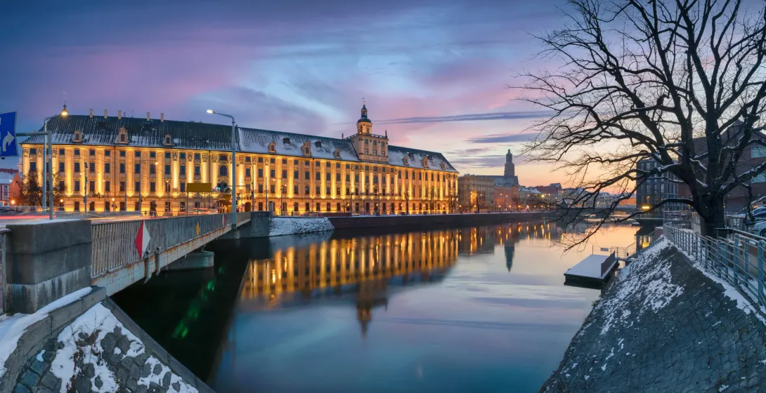 Illuminated University of Wrocław building and bridge over the Odra River at dusk, with snow on banks.