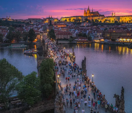 Tourists stroll across Charles Bridge in Prague at sunset, with Prague Castle majestically lit in the background.