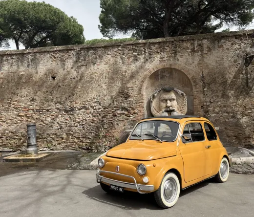 A yellow Fiat 500 parked near a Roman wall with a unique fountain.