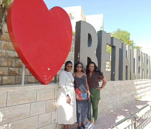 Three women pose happily in Bethlehem, Palestine, in front of a large red heart and the city's name.