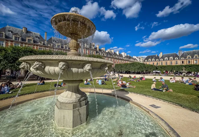 A picturesque fountain in Place des Vosges, Le Marais, Paris, surrounded by people enjoying a sunny day.
