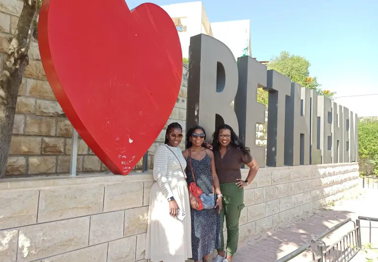 Three women pose happily in Bethlehem, Palestine, in front of a large red heart and the city's name.