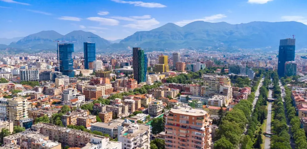 Aerial view of Tirana, Albania, showcasing modern skyscrapers and traditional buildings against a mountain backdrop.