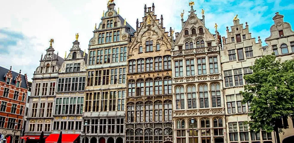 Elaborate historic guildhalls with stepped gables and golden statues line a cobblestone market square in Antwerp. People walk