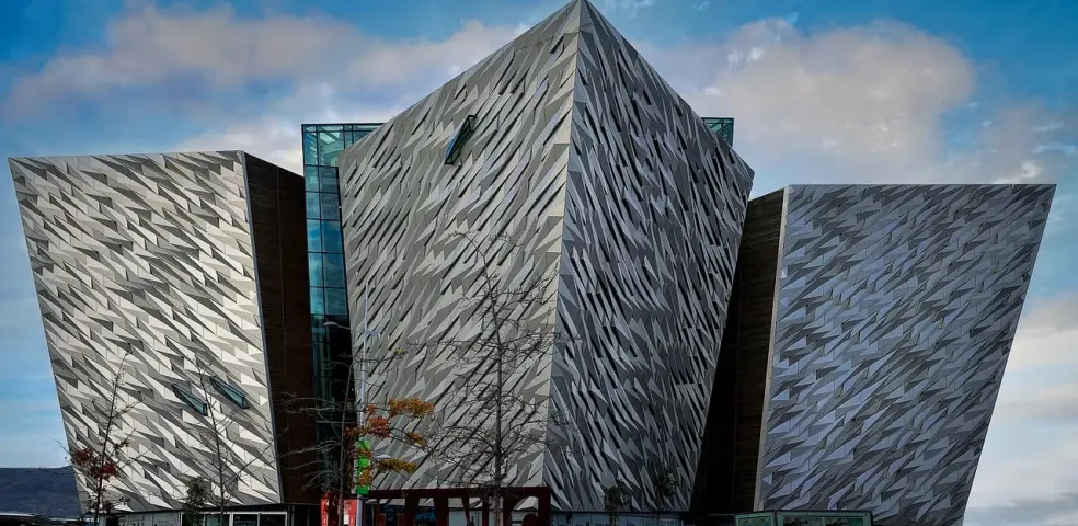 The Titanic Belfast building with its angular, faceted silver panels under a partly cloudy sky, with bare trees in the foregr