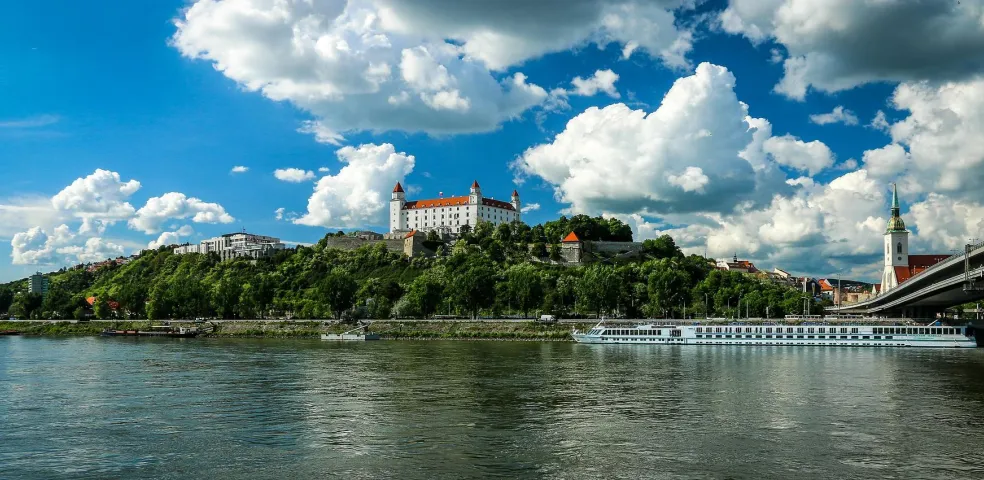 Bratislava Castle on a green hill overlooking the Danube River, with a cruise ship, bridge, and St. Martin's Cathedral spire.