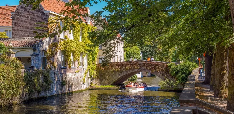 A tour boat with passengers on a canal in Bruges, passing under a stone arch bridge next to an ivy-covered building.