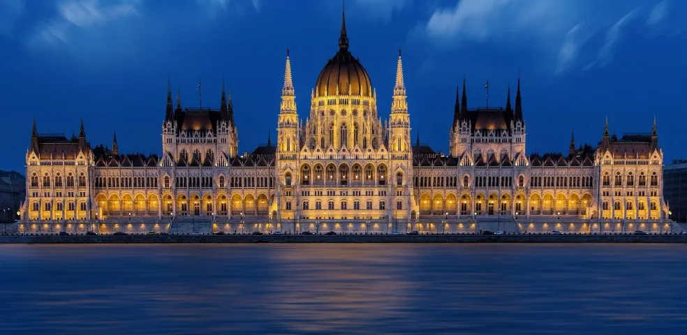 The illuminated Hungarian Parliament Building at night, viewed across the dark blue Danube River under a cloudy sky.