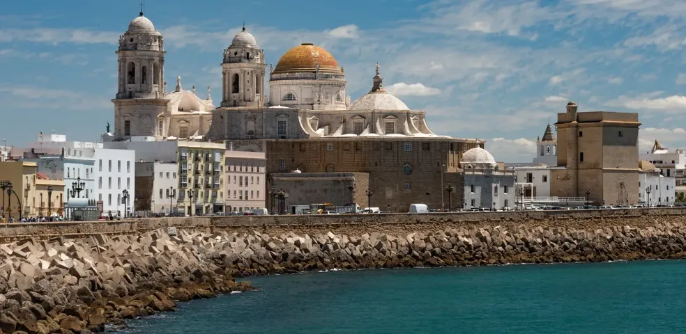 View of Cadiz Cathedral and city skyline from the sea, with a rocky breakwater in the foreground under a blue sky.