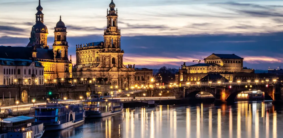 Dresden city skyline at dusk with illuminated historic buildings along the Elbe River, boats docked, and reflections on the w
