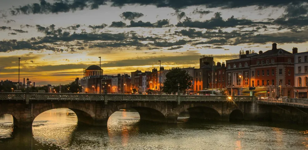 Stone arch bridge over a river in Dublin at sunset, with city buildings lit up on the far bank and a cloudy sky.