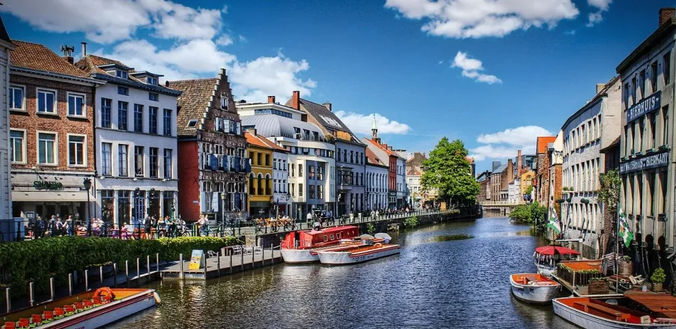Historic buildings with gabled roofs line a sunny canal with several tour boats and people walking on the banks.