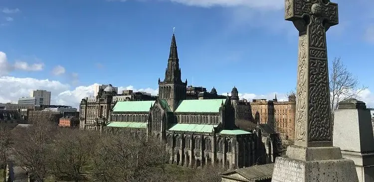 Historic Glasgow Cathedral with green roof and spire, viewed from the Necropolis, showing an ornate Celtic cross in the foreg