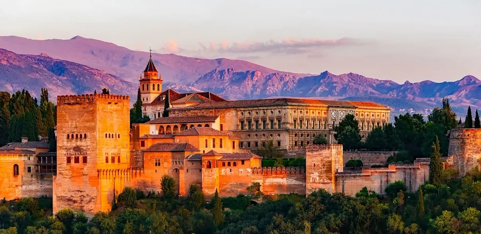 Alhambra palace complex with fortified walls and towers at sunset, mountains in the background, surrounded by green trees.