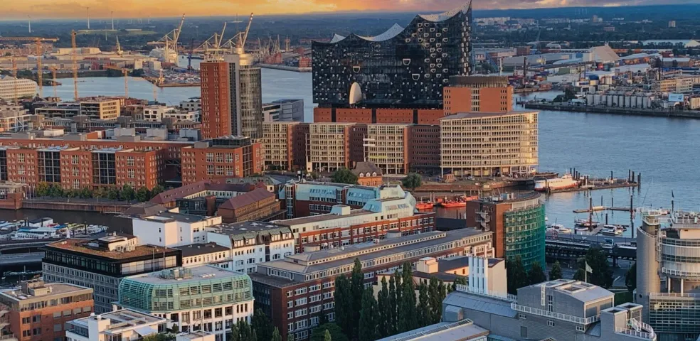 Aerial view of Hamburg harbor at sunset, featuring the Elbphilharmonie, brick buildings, and industrial cranes by the water.