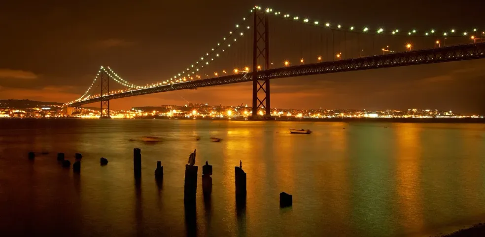 25 de Abril Bridge spanning a river at night, illuminated with city lights reflecting on the water. Wooden posts in foregroun