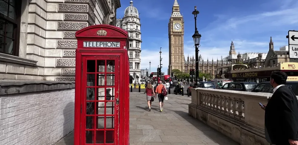 A classic red telephone booth on a London sidewalk with Big Ben and the Houses of Parliament in the background under a blue s