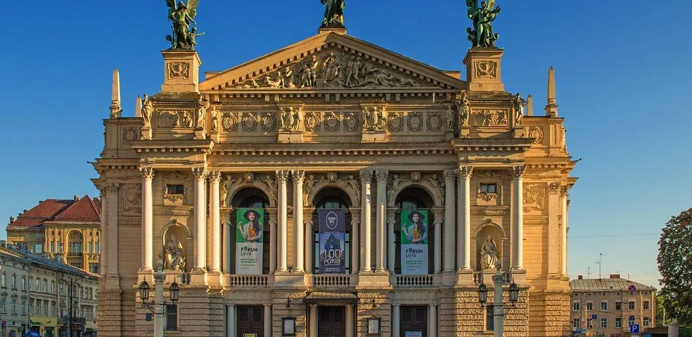 Facade of the Lviv Opera House with columns, sculptures on the roof, and banners. Clear blue sky above. People walk in the fo
