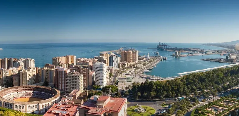 Panoramic view of Malaga cityscape with La Malagueta bullring, the port, and the Mediterranean Sea under a blue sky.