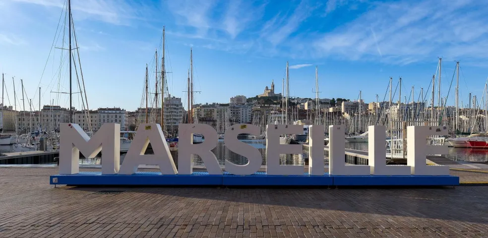 Large MARSEILLE letters on a blue base at the Vieux-Port, with sailboats and buildings. Notre-Dame de la Garde visible on the