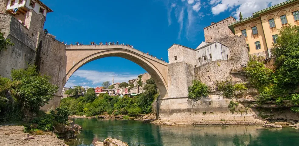 Historic Stari Most bridge in Mostar, Bosnia and Herzegovina, spanning a green river with ancient stone buildings and lush ba
