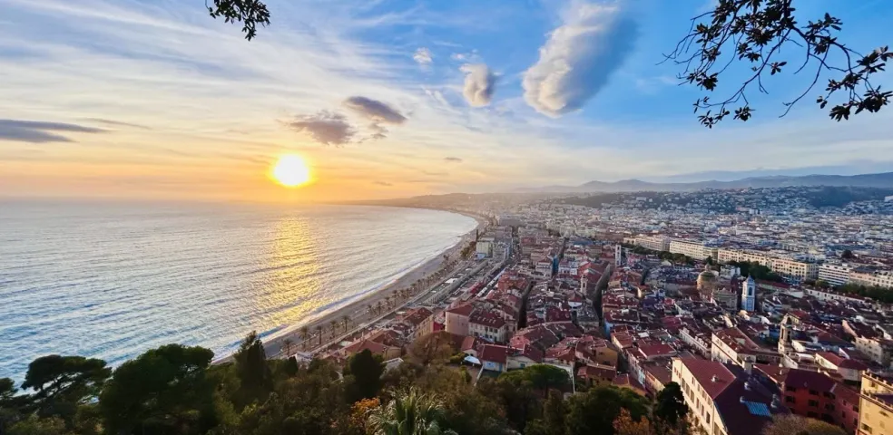 Panoramic view of Nice coastline at sunset, showing the sun reflecting on the sea, a long beach, promenade, and the city buil