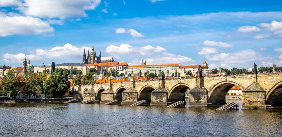 View of Charles Bridge with statues and arches spanning a river, Prague Castle on the distant left bank under a blue sky with