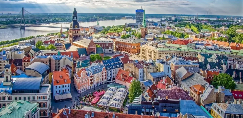 Aerial view of Riga Old Town with colorful buildings, St. Peter's Church spire, Daugava River, and Vansu Bridge under a cloud