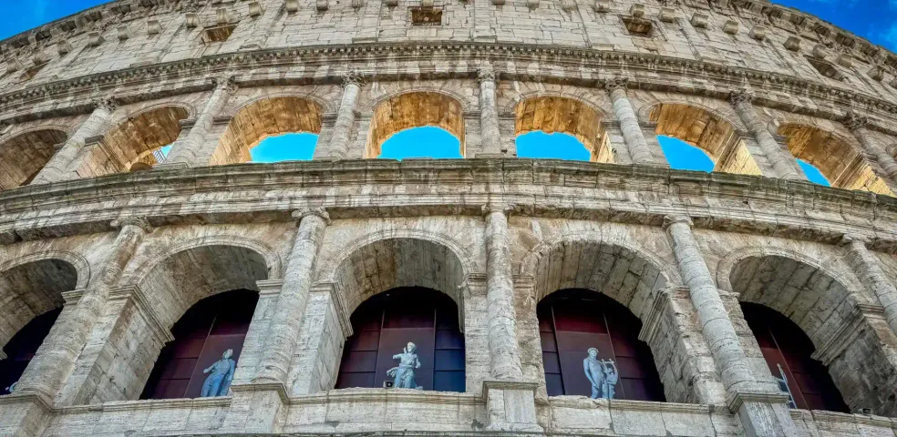 Exterior view of the Colosseum in Rome, showing multiple tiers of stone arches, columns, and statues in lower arches, against