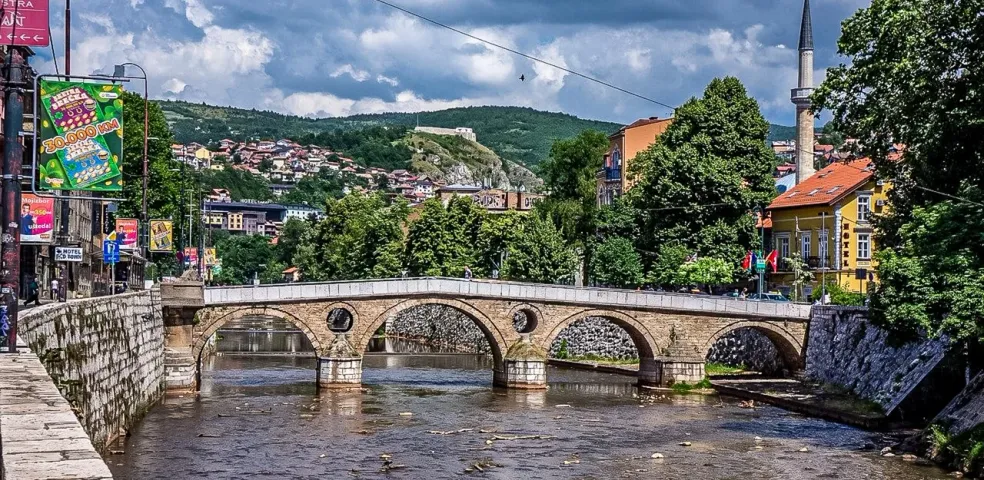 Historic stone arch bridge over a river in Sarajevo, with a minaret, buildings, and green hills under a cloudy sky.