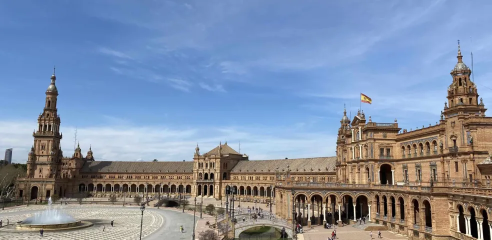 Large ornate building with arcades, a central canal with bridges, patterned plaza, and fountain under a blue sky in Seville.