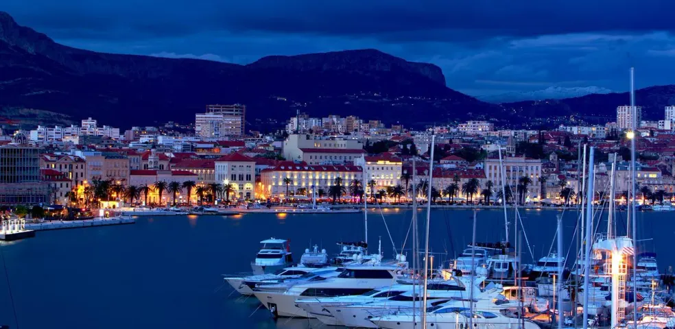 Harbor at twilight with numerous yachts and boats, illuminated waterfront buildings, palm trees, and mountains under a dark b