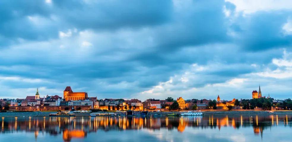 Toruń skyline at dusk, with historical buildings illuminated along the riverbank, reflected in the calm water under a cloudy