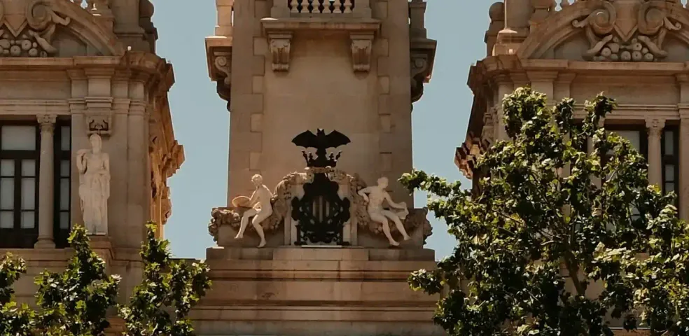 Ornate clock tower of Valencia City Hall with a large clock face, statues, flags, and green trees in foreground under a clear