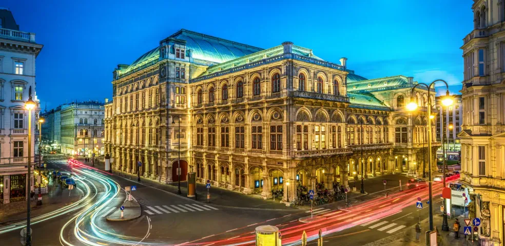 Illuminated Vienna State Opera building at twilight, with light trails from cars on a street crossing and a crescent moon in