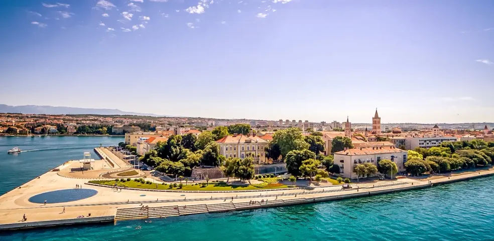 Aerial view of Zadar waterfront with the Sea Organ, old town buildings, clear blue sea, and distant mountains under a sunny s