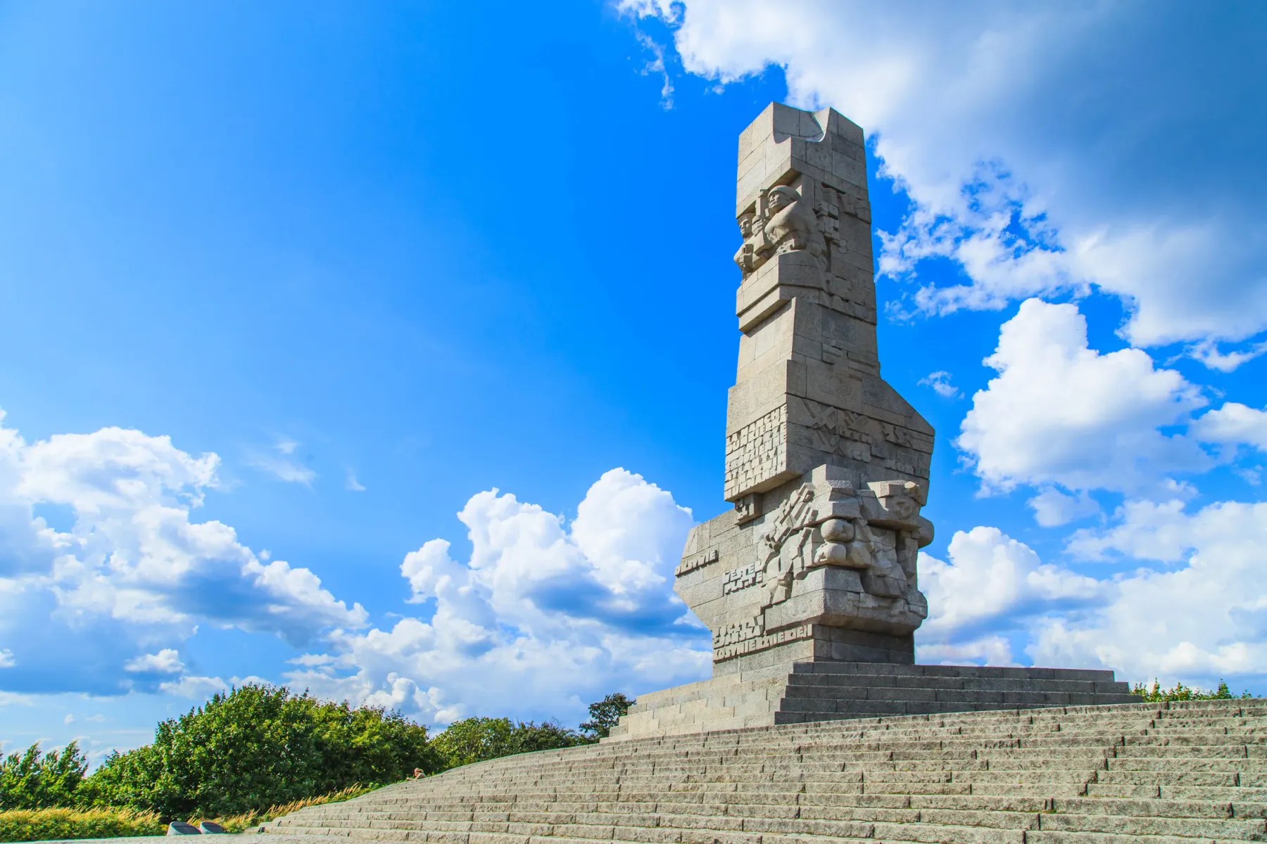 Tall concrete Westerplatte Monument with carved figures, on a wide stepped base, against a bright blue sky with white clouds.