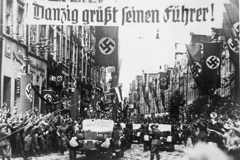 Black and white historical image of a street in Gdansk (Danzig) during a Nazi parade with crowds, vehicles, swastika flags...