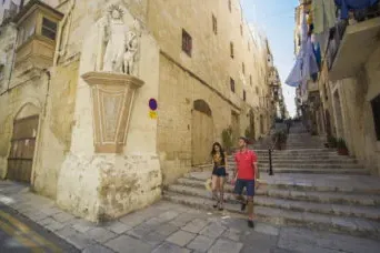 Two people walk down historic stone steps on a narrow street in Valletta, flanked by old buildings and a statue on a corner.