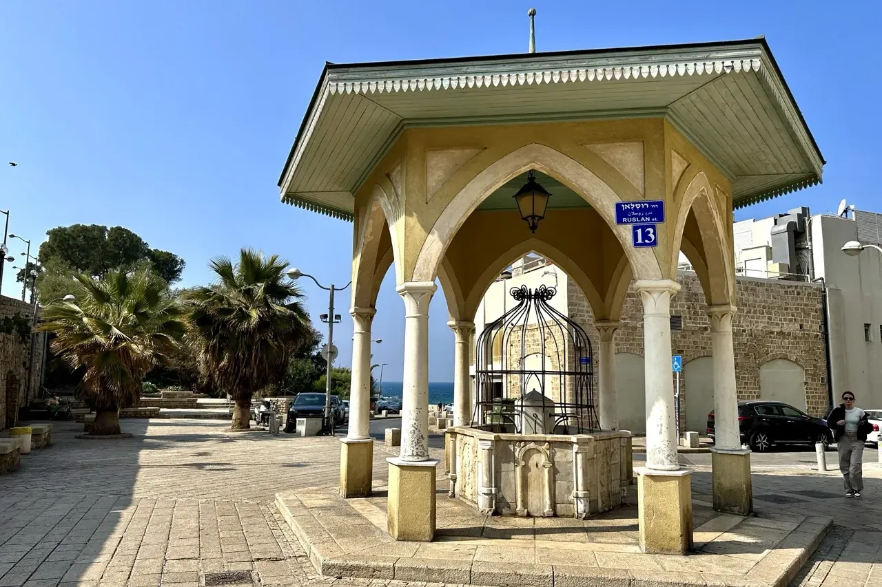Ornate yellow gazebo with a light green roof, sheltering a stone fountain on a paved square with palm trees and a street s...