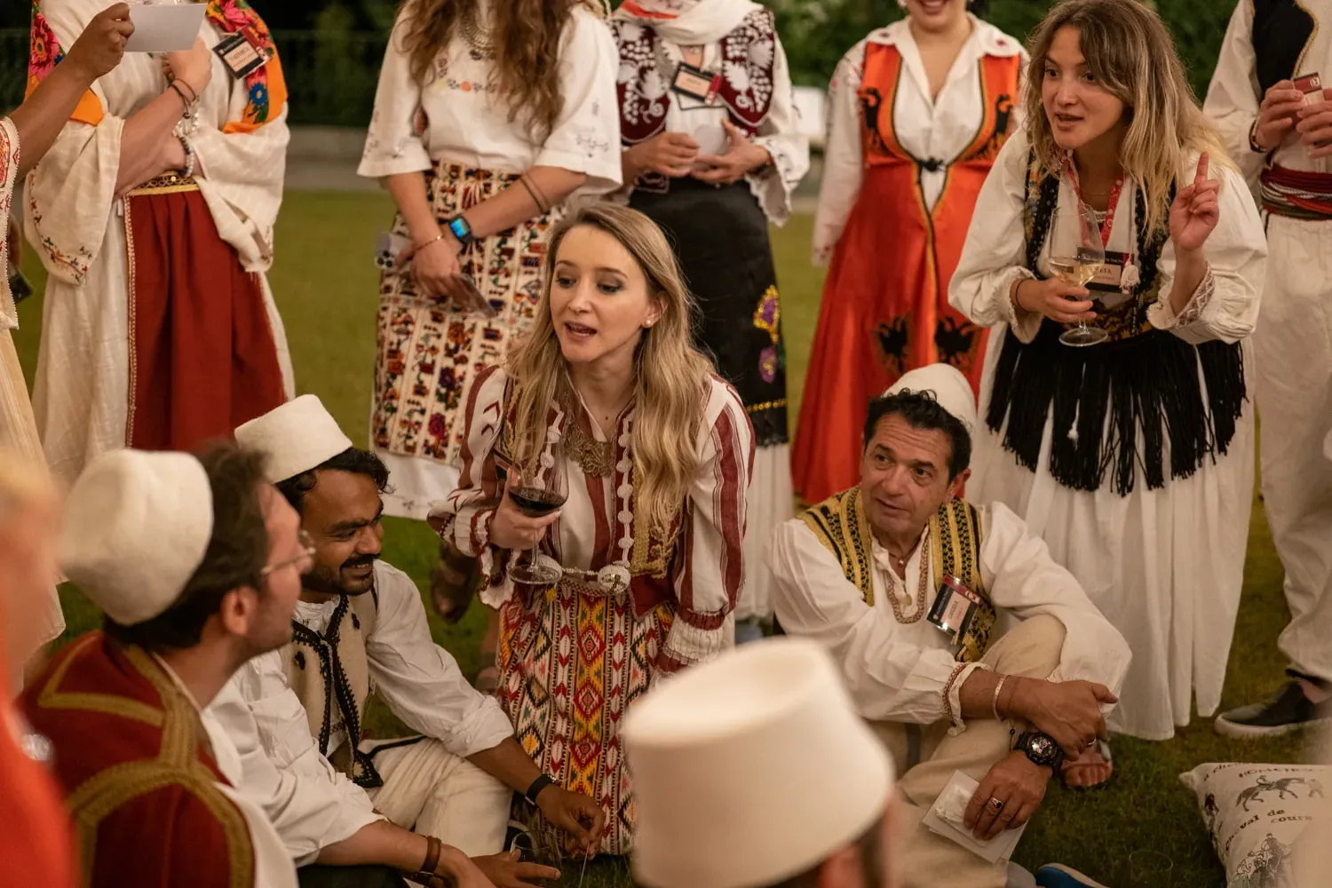 People in various traditional Albanian clothing with white Qeleshe hats, holding drinks, gathered on grass at night.