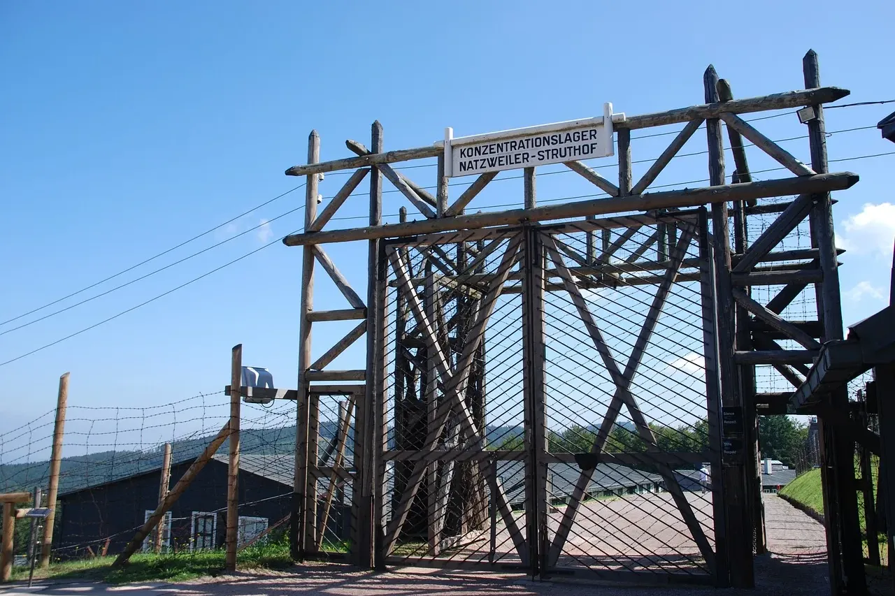 Large wooden gate with barbed wire fence and a sign reading 'KONZENTRATIONSLAGER NATZWEILER-STRUTHOF' under a clear blue sky.