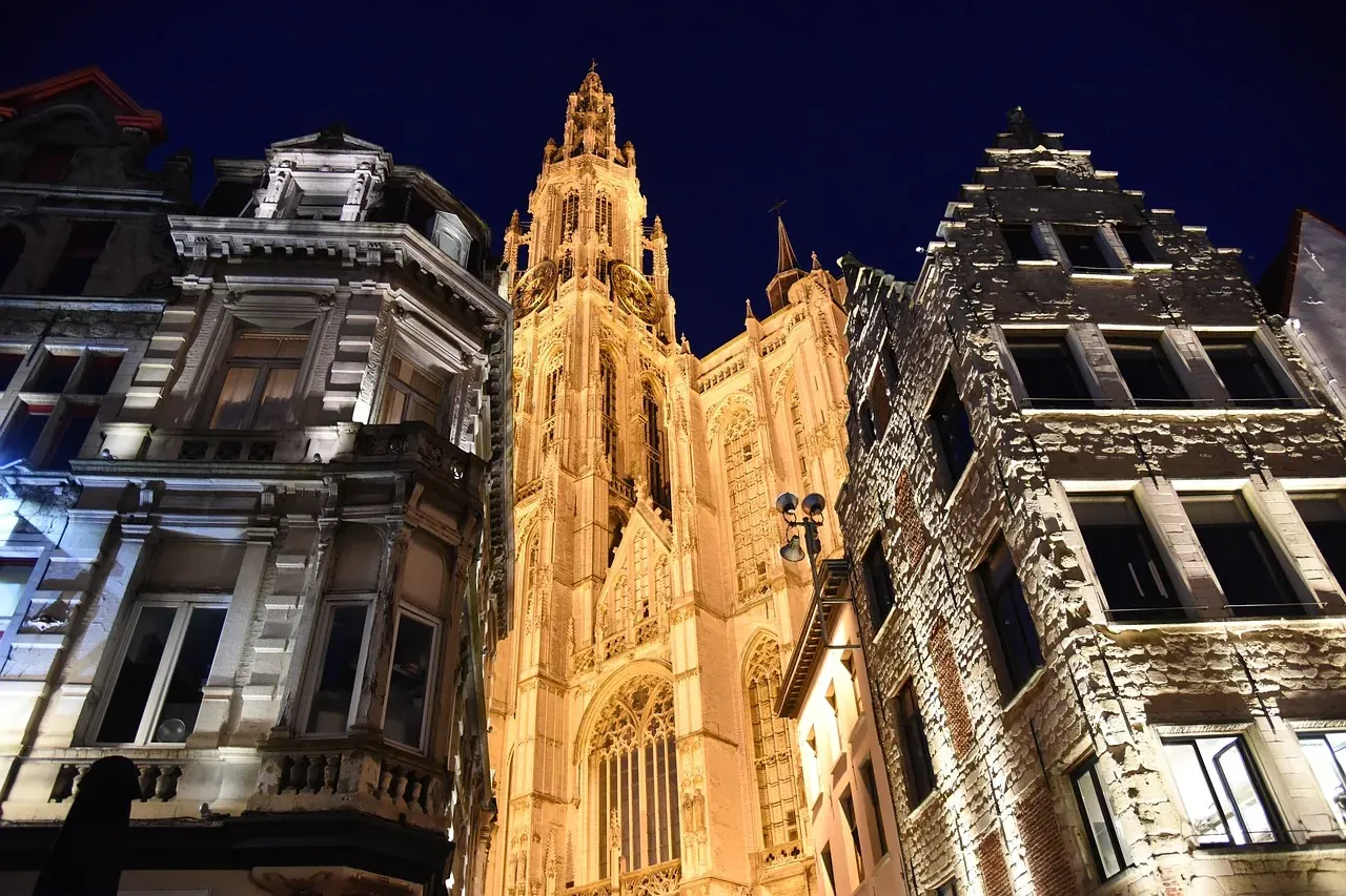 Illuminated Gothic cathedral tower at night, flanked by historic European buildings under a dark blue sky.