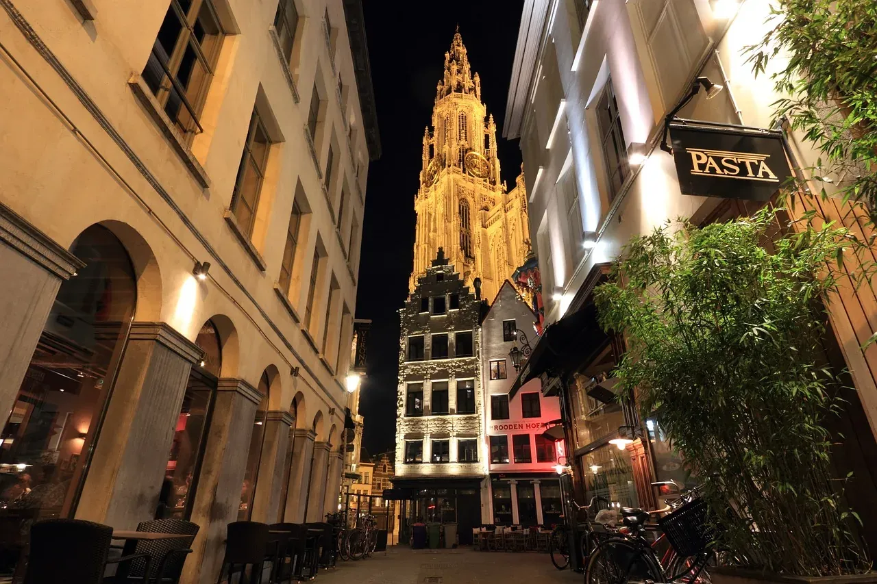 The illuminated tower of Antwerp Cathedral of Our Lady at night, seen from a narrow cobblestone street with buildings.