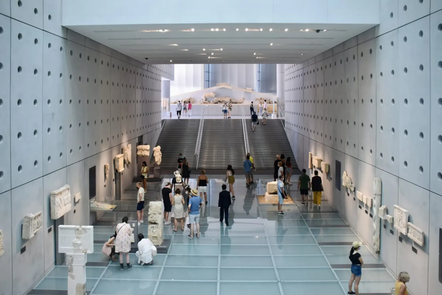 Modern museum interior with ancient sculptures on concrete walls, glass floor, and visitors. A grand staircase leads to a ...