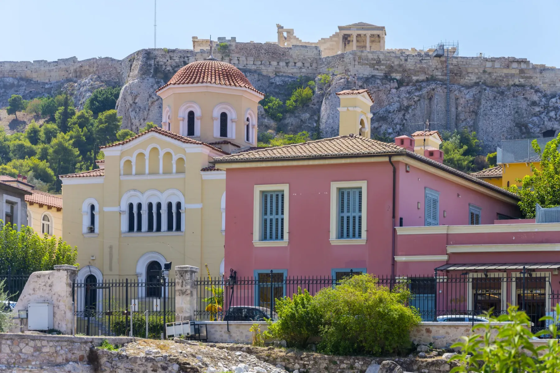 Yellow domed church and a pink building in Athens, with the rocky Acropolis hill and Parthenon ruins in the background.