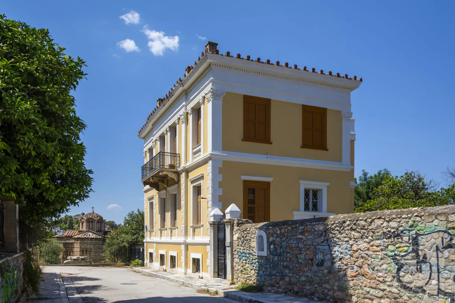 Yellow neoclassical building with white trim, brown shutters, and a balcony on a street. A stone wall with graffiti and a ...