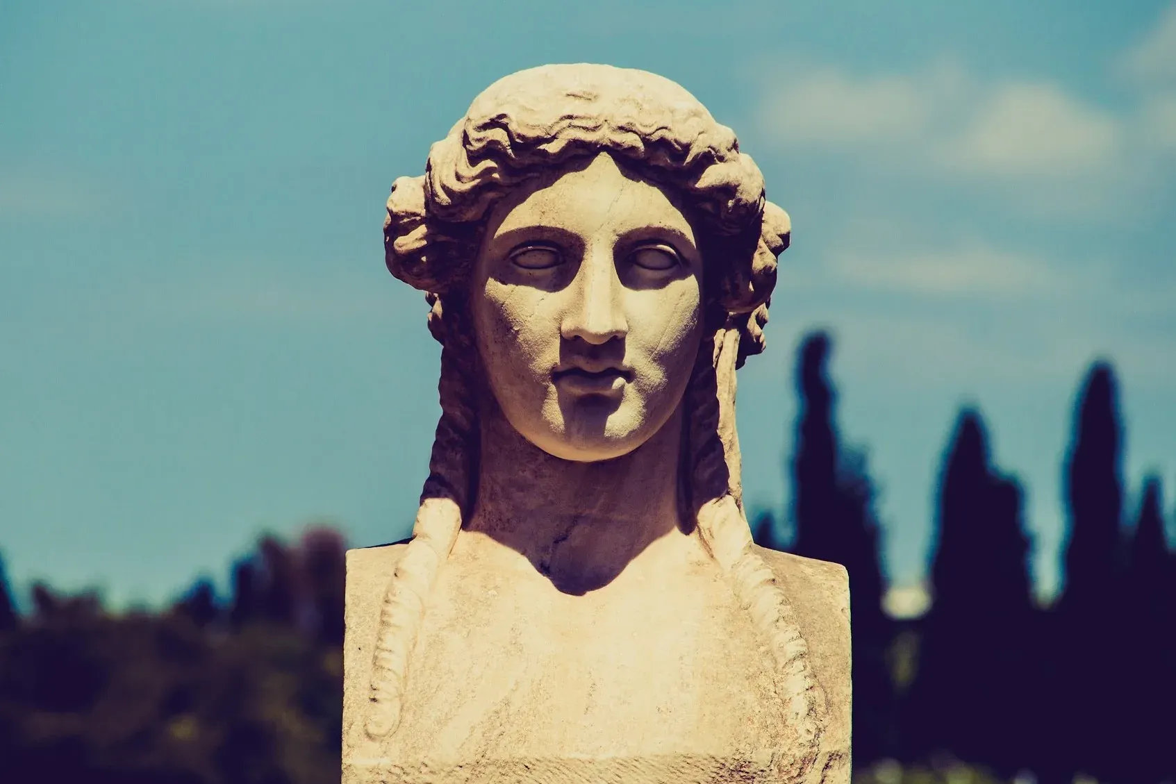 Close-up of an ancient Greek marble bust of a woman with braided hair, against a clear blue sky.