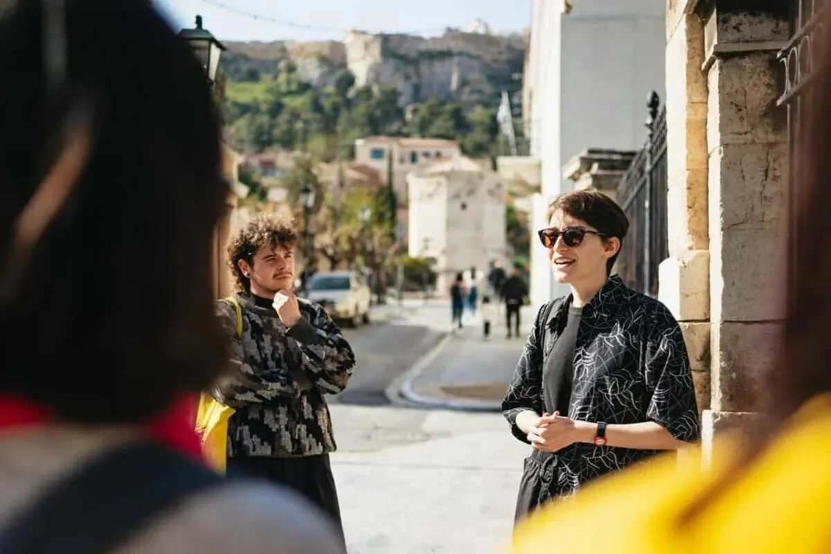 A smiling tour guide in sunglasses talks to a small group of tourists on a sunny street in Athens, with the Acropolis in t...